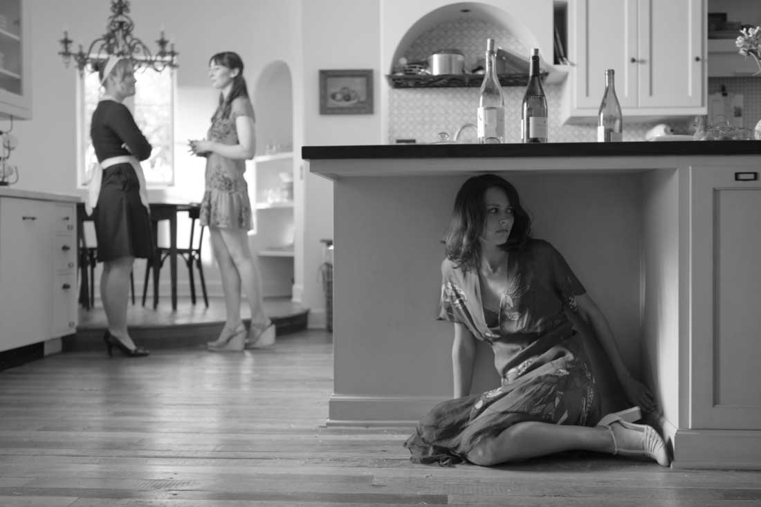 Amy Acker (under counter) hides from her friends’ conversation about her love life in Much Ado About Nothing.