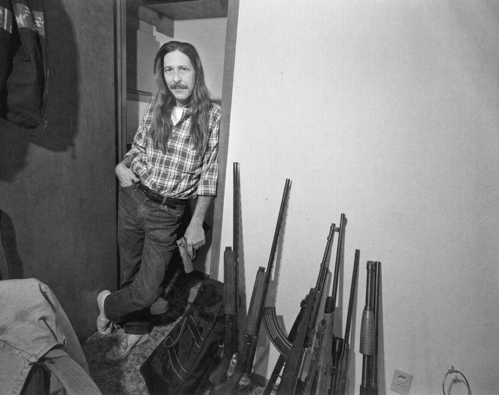 A drug dealer or, as Byrd IV describes him, an “addictive substance vendor,” poses with his tools of the trade –– a handgun and an array of shotguns and rifles. Check out the curved clip on the middle rifle and the hair and mustache (and tucked-in shirt) of the dapper dealer.